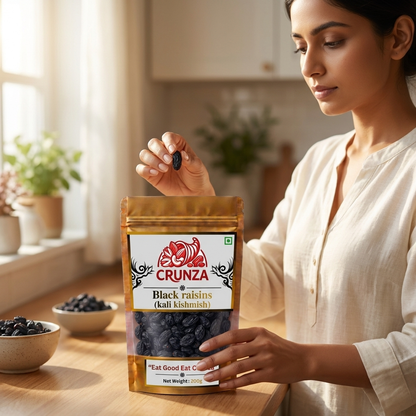A woman in a white blouse holds a bag of CRUNZA Black raisins (kali kishmish) on a wooden table in a bright kitchen setting, with bowls of raisins nearby.