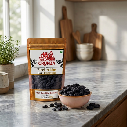 A stand-up pouch of CRUNZA Black raisins (kali kismiish) stands on a marble kitchen countertop next to a small bowl filled with dark raisins and a few scattered raisins.