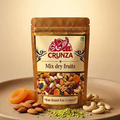A stand-up pouch of CRUNZA Mix dry fruits on a beige surface, surrounded by almonds, cashews, dried apricots, and green pistachio pieces. The pouch is brown with a cream label featuring the red CRUNZA logo.