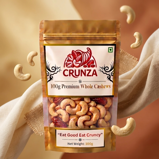 A stand-up pouch of CRUNZA 100g Premium Whole Cashews centered against a warm beige and brown background with draped fabric and floating cashews. The pouch has a metallic gold top and base, a white front label with red and black branding, and a clear window showing the cashews inside.
