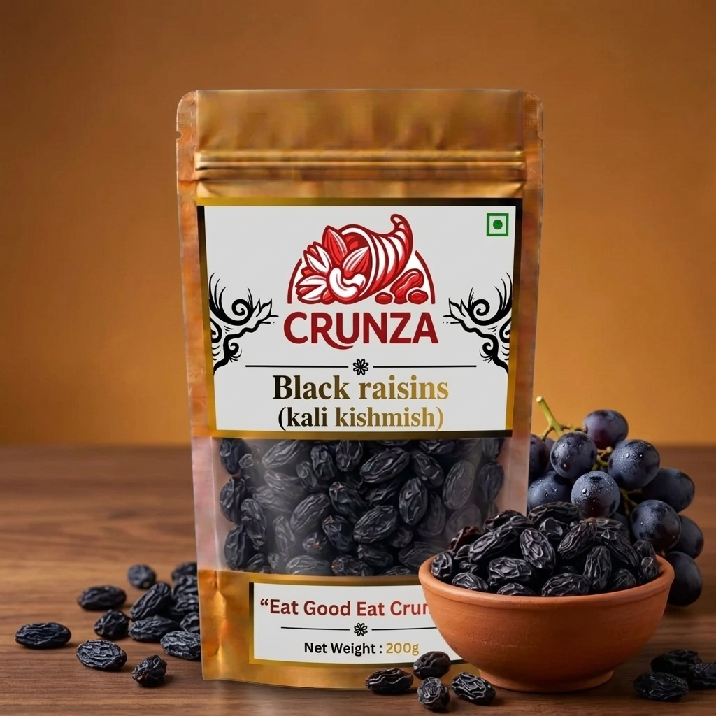 A stand-up pouch of CRUNZA Black raisins (kali kishmish) displayed on a wooden surface with a bowl of fresh dark grapes and scattered raisins against a warm brown background.