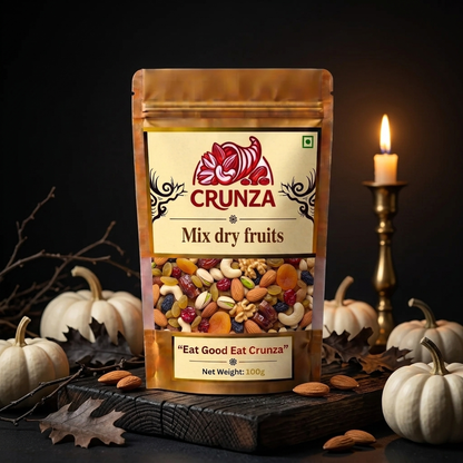 A 100g stand-up pouch of CRUNZA Mix Dry Fruits, styled on a dark wooden board with white pumpkins, almonds, and a burning candle in the background.