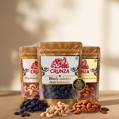 Three stand-up pouches of CRUNZA nuts and dried fruits arranged together on a beige surface with loose almonds, cashews, and black raisins in front. The center pouch is labeled Black raisins (kali kishmish), with additional labels showing California Almonds and Premium Cashews behind it.