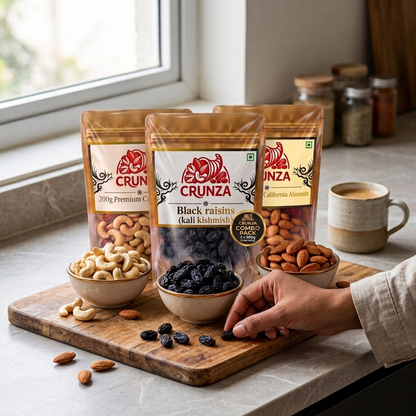Three stand-up pouches of CRUNZA nuts and dried fruits arranged on a wooden board on a kitchen counter, with bowls of cashews, almonds, and black raisins, plus a hand reaching toward the center pouch.